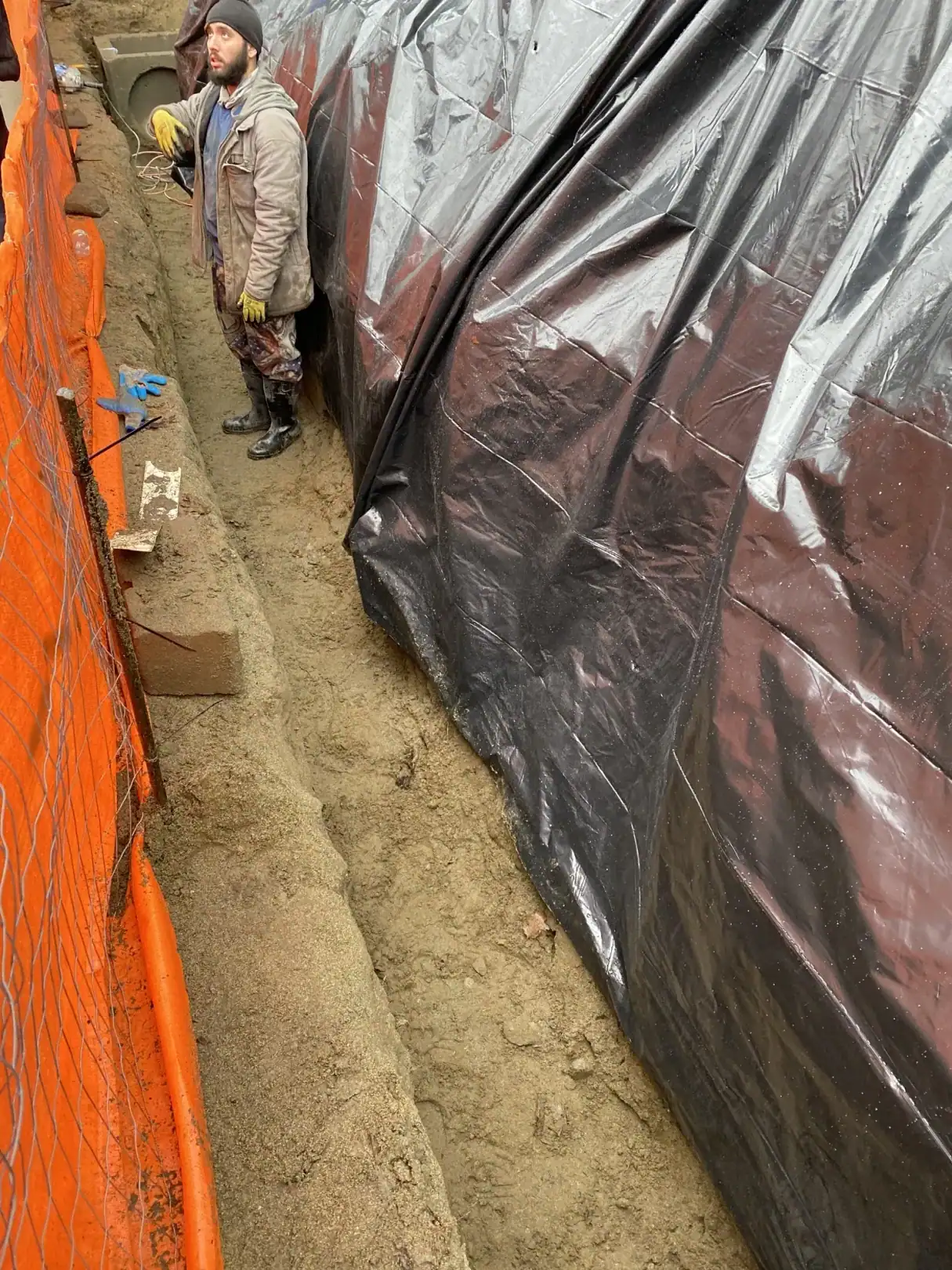 Worker applying waterproofing membrane to the block retaining wall behind orange safety fence