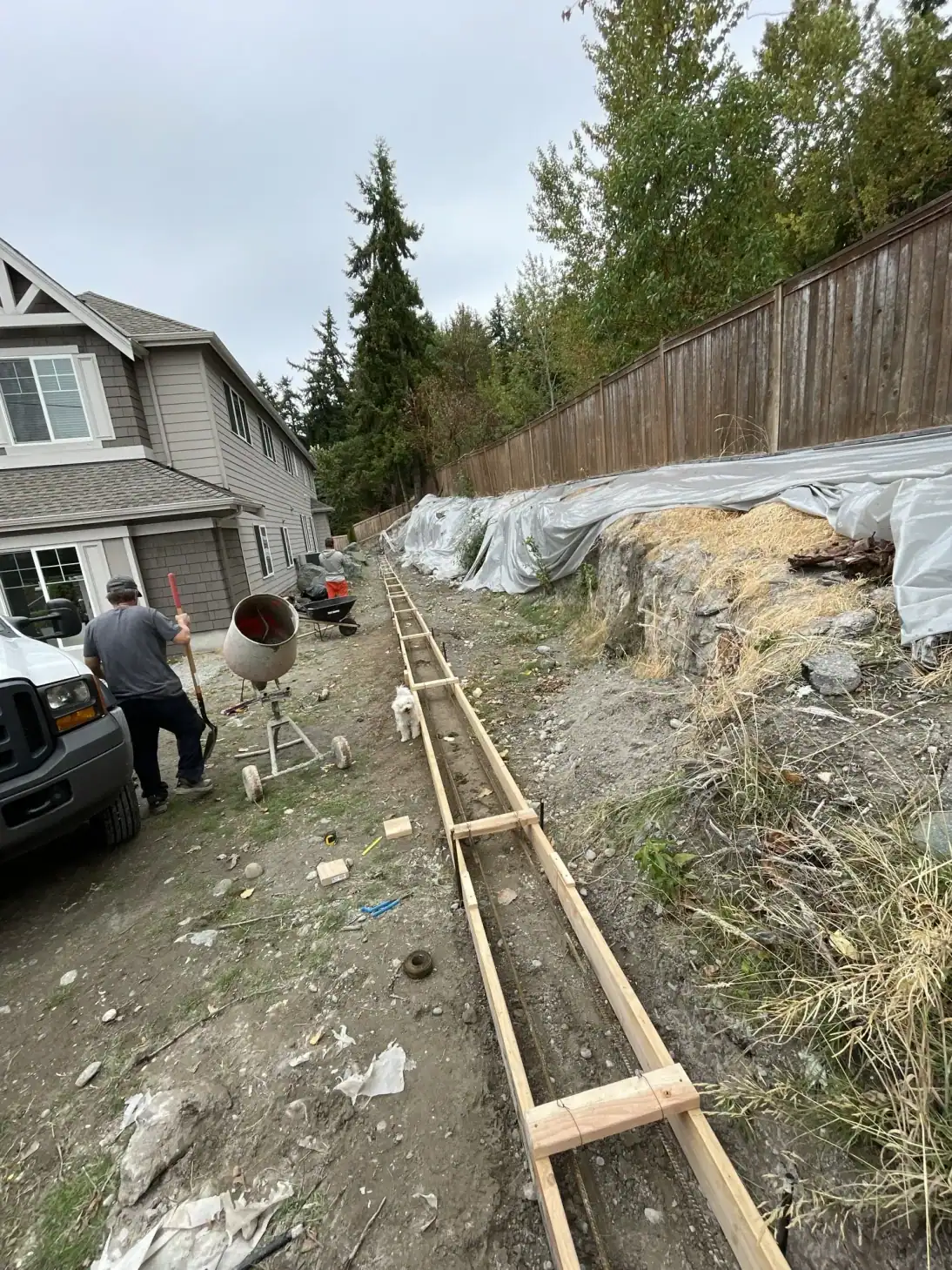 Footing forms running toward the house with a concrete mixer and sheeting on the bank