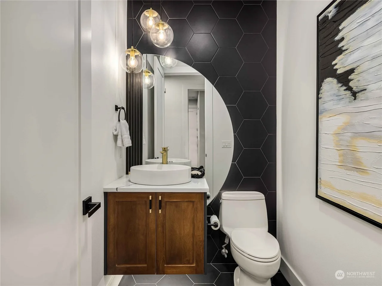 Powder room with a black hexagon tile wall, round mirror, walnut vanity, vessel sink, and gold sconces