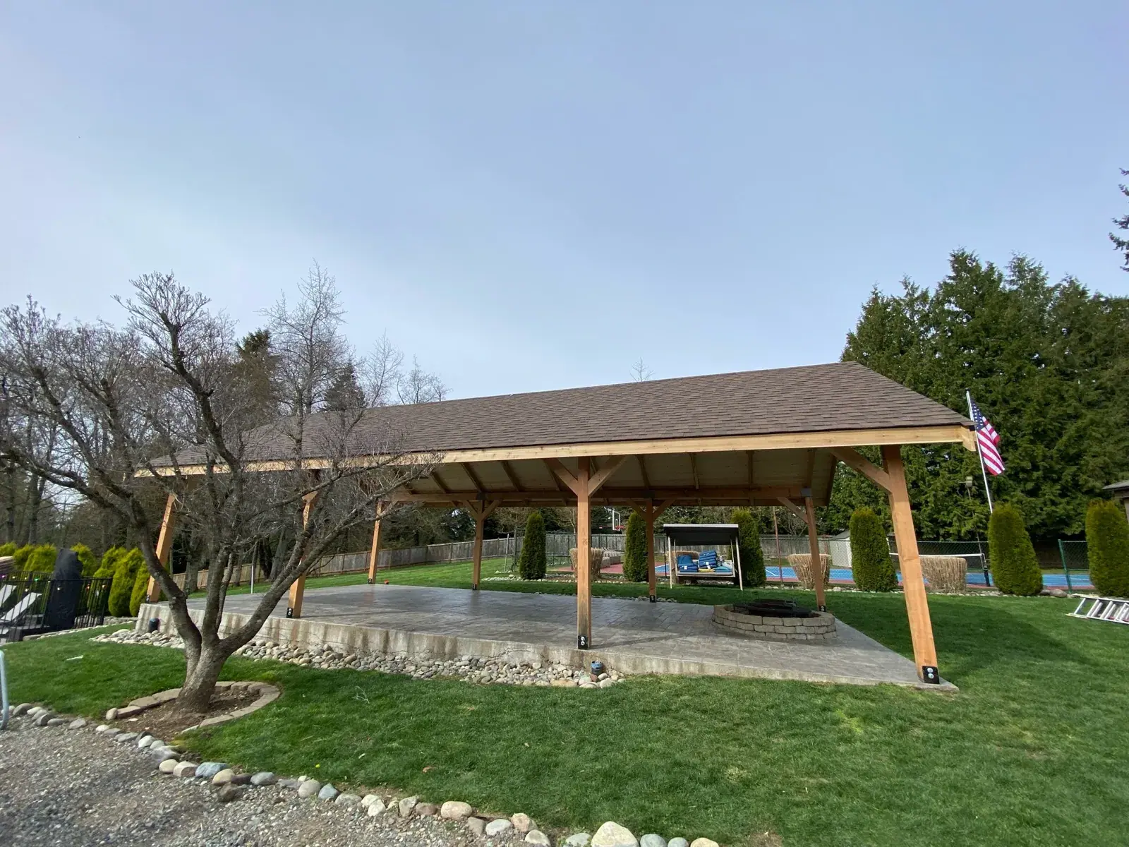 Finished pavilion with a brown architectural shingle roof on the lawn beside a flag