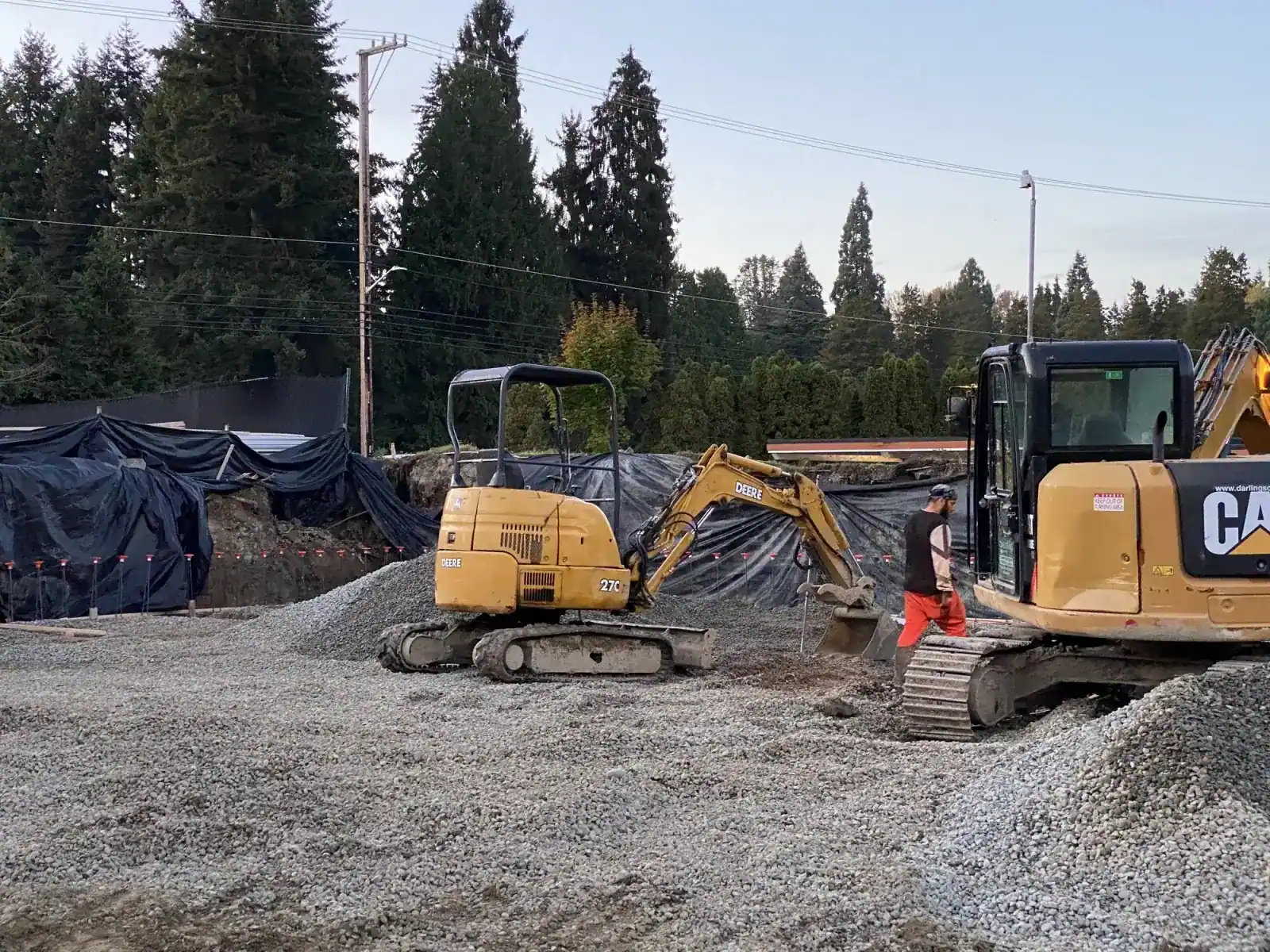 Cat mini-excavators beside gravel piles and a black-plastic-covered soil stockpile