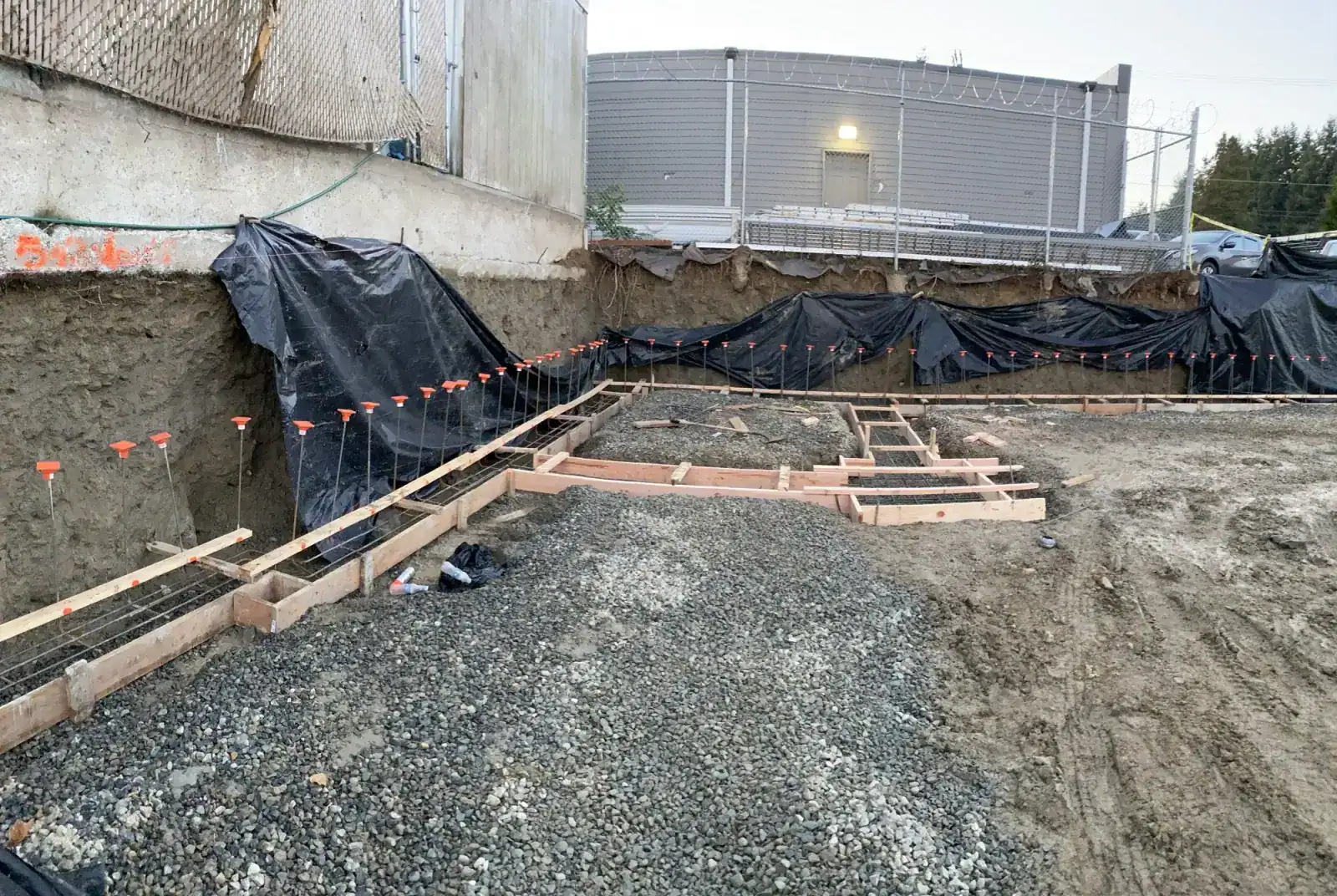 Formed footings with rebar on gravel, orange marker flags, and black plastic on the cut bank