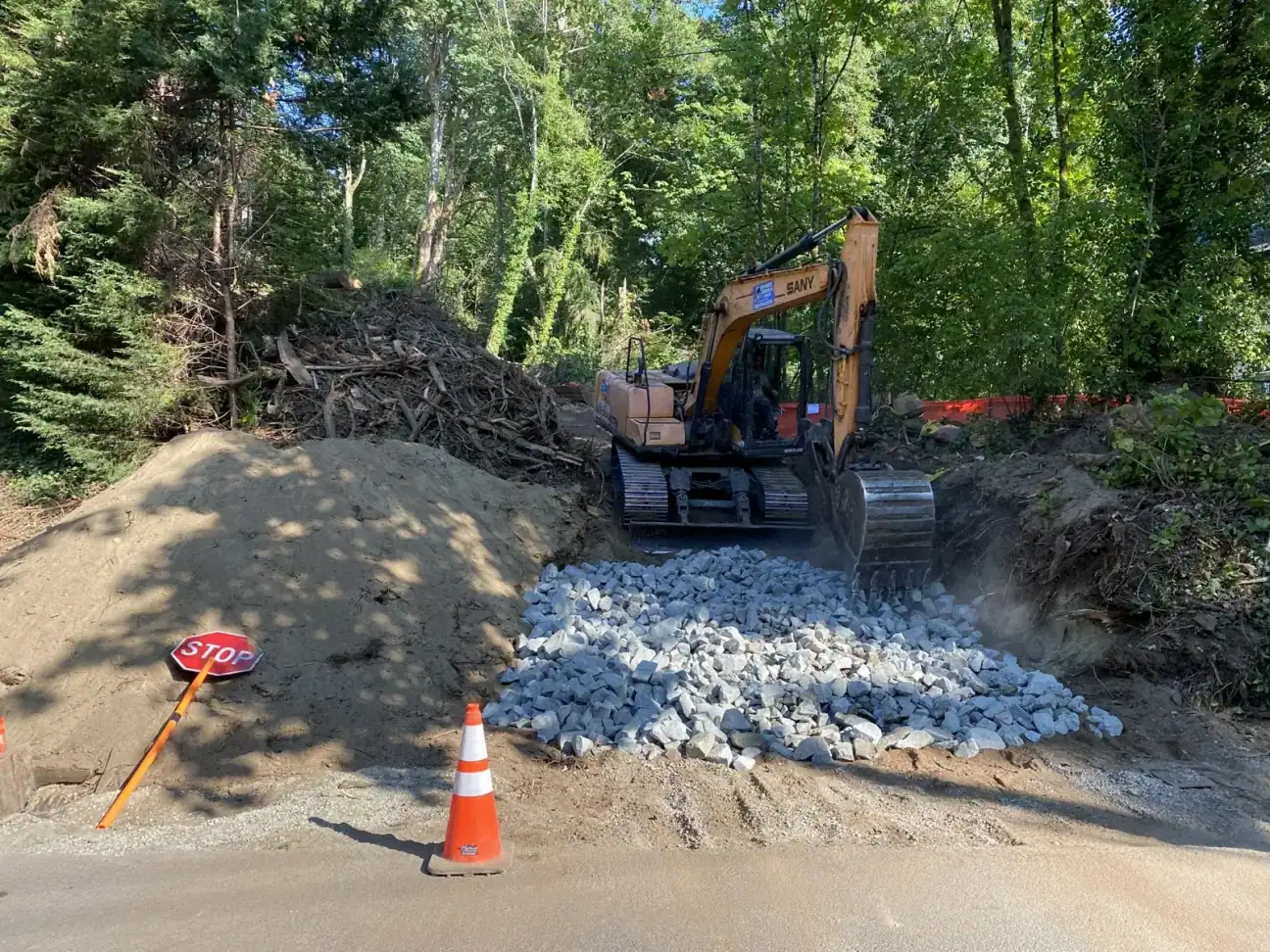 Excavator spreading quarry rock at the site entrance beside a traffic cone