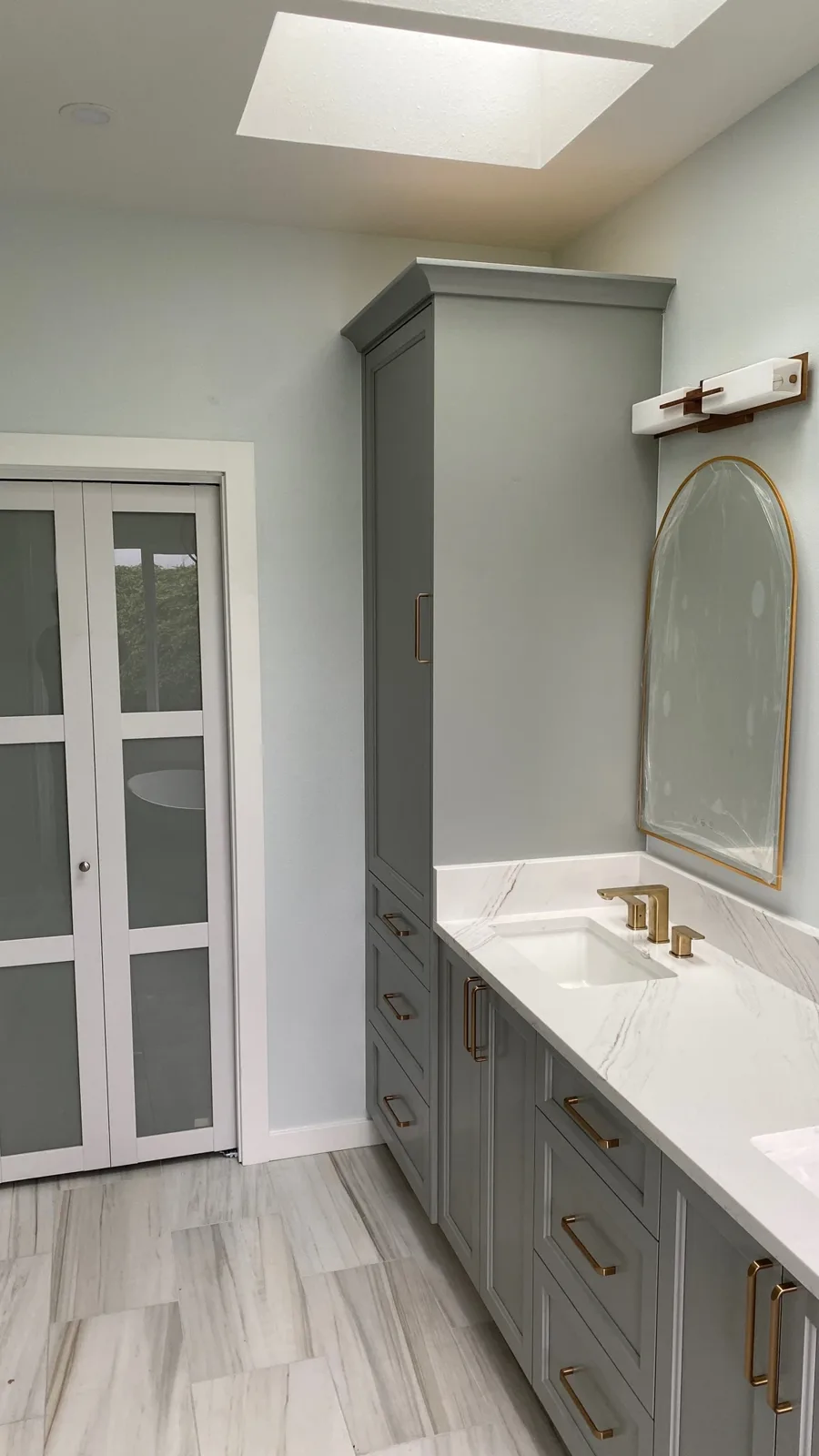 Gray double vanity with arched mirrors, gold sconces, and frosted bifold closet doors under a skylight