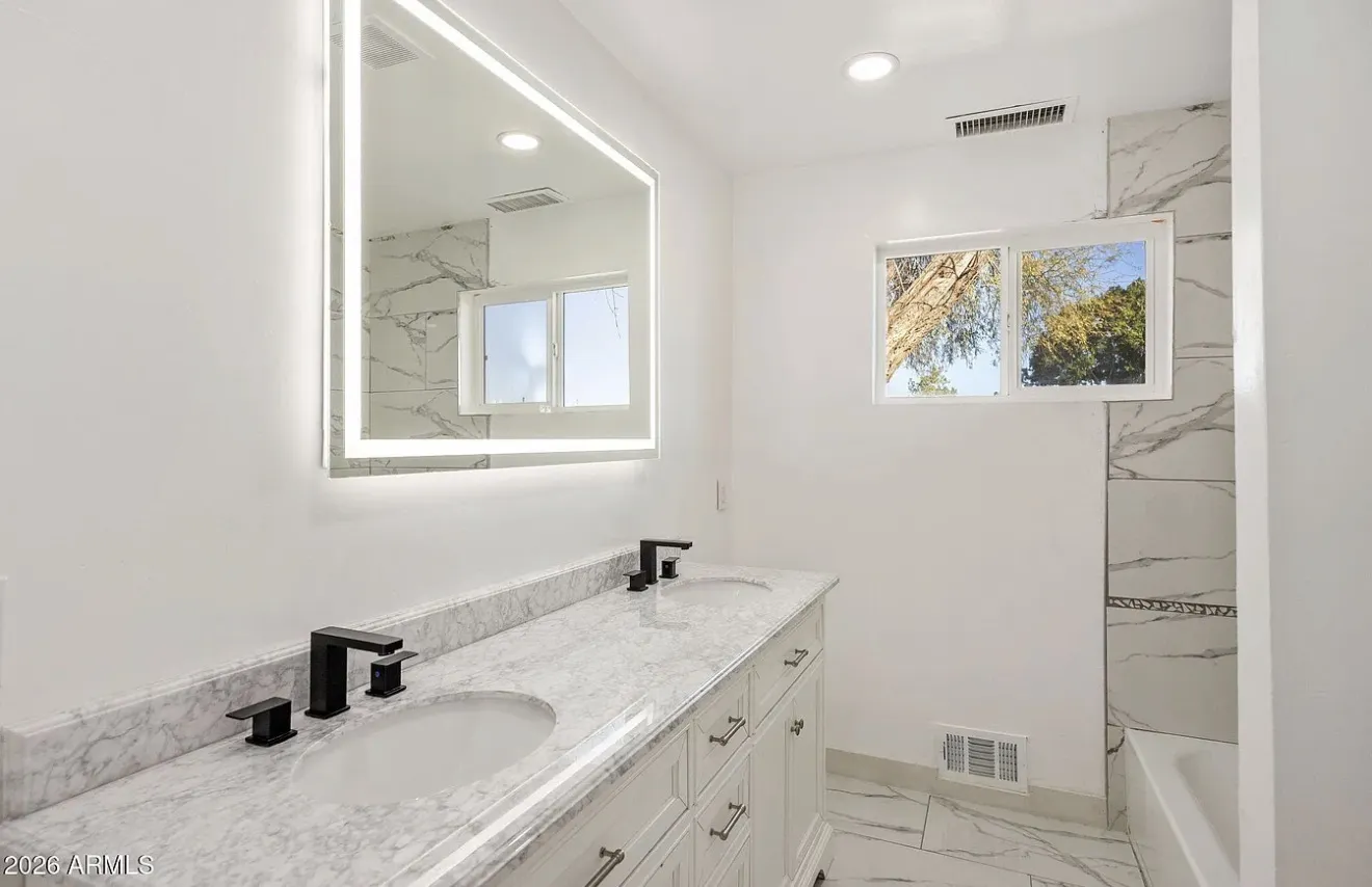 Bathroom with a double vanity, marble counter, LED mirror, and marble-tile tub surround